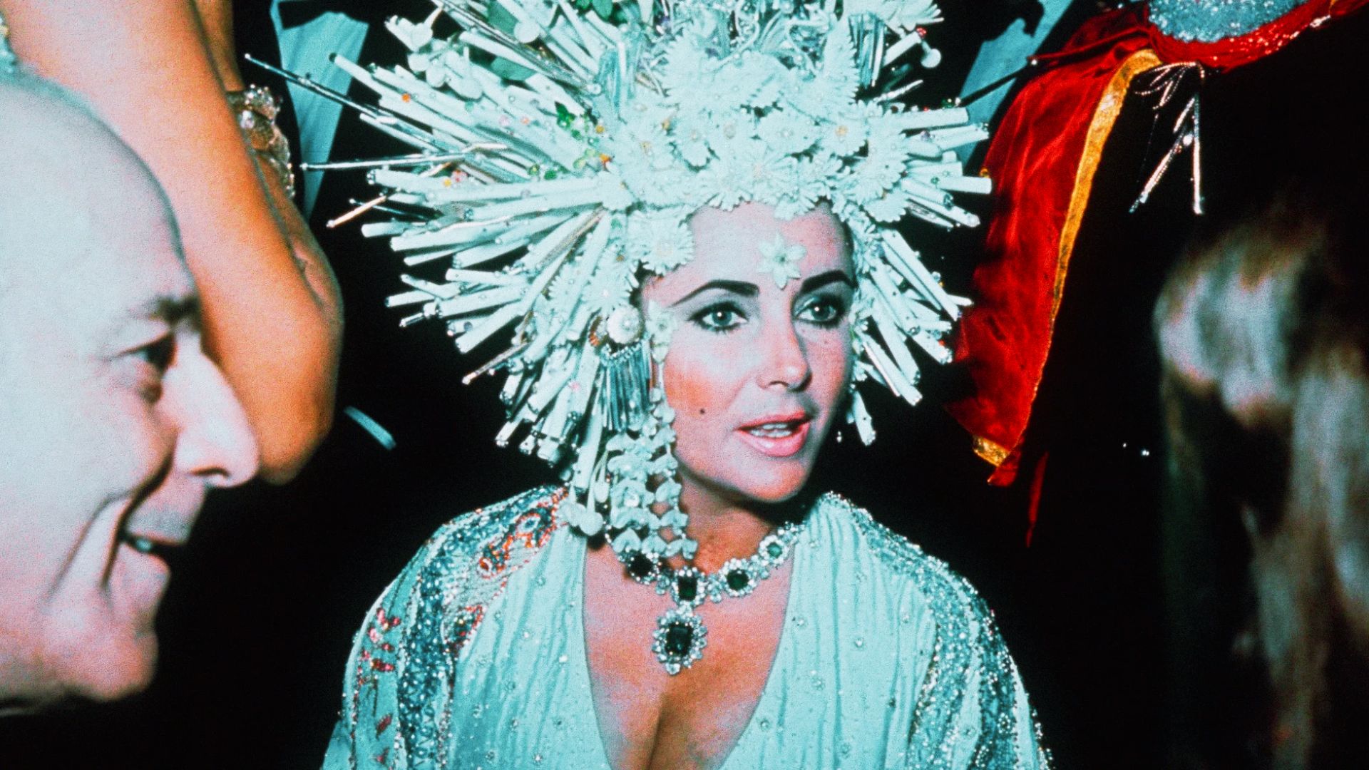 Elizabeth Taylor at a gala in an ornate white headdress and emerald Bulgari necklace.