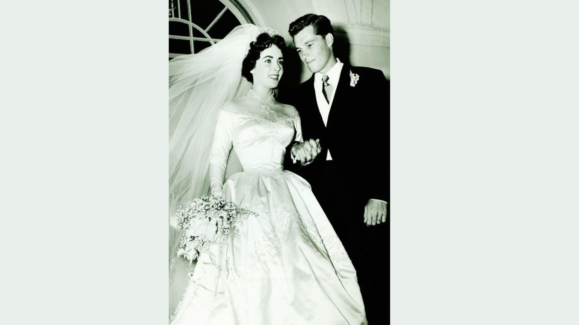 Black-and-white photo of Elizabeth Taylor in the same 1950 wedding dress, gazing at her groom under an arched doorway.