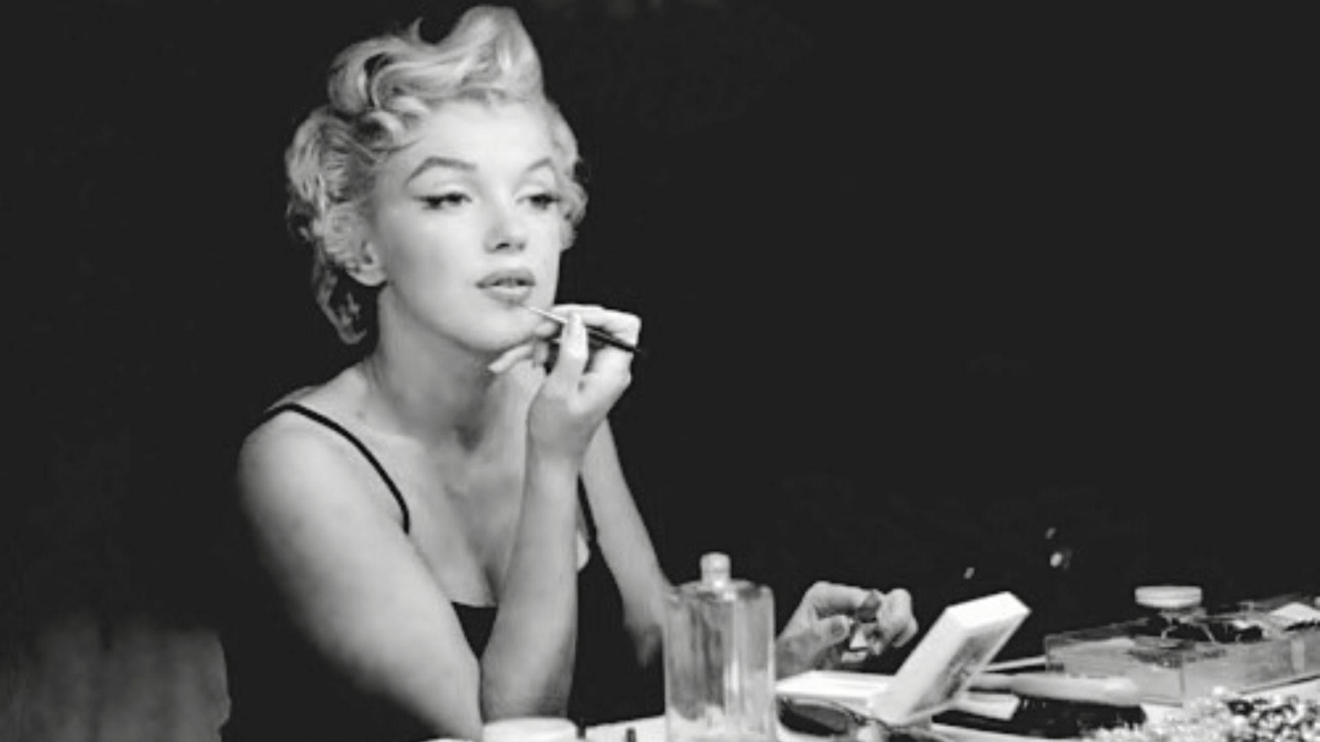 Black and white photo of Marilyn Monroe applying makeup at vanity with perfume bottle, linked to her Chanel No 5 legacy