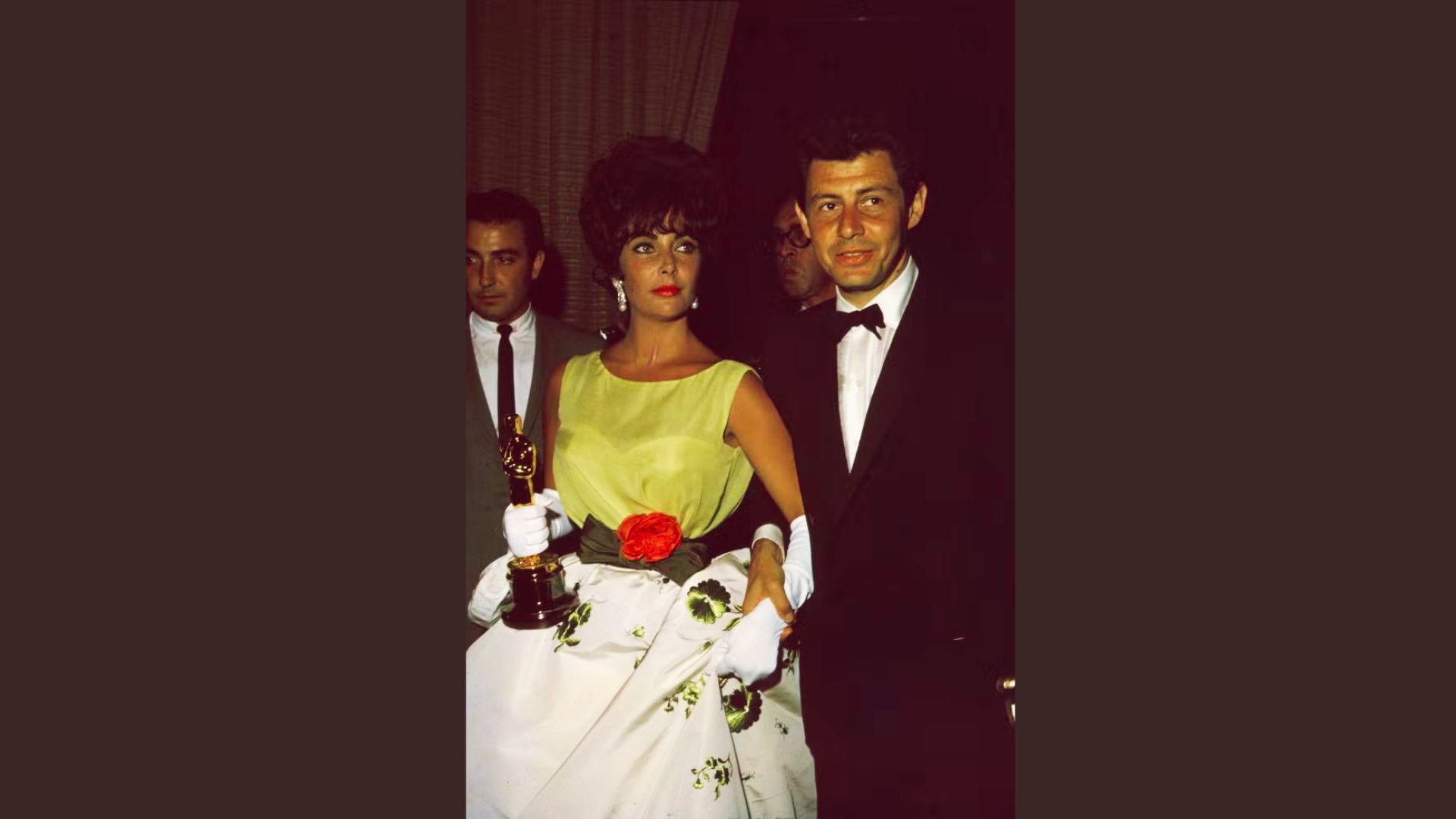 Elizabeth Taylor holds her Oscar in a yellow bodice gown with floral skirt, white gloves, and a red rose at the waist.