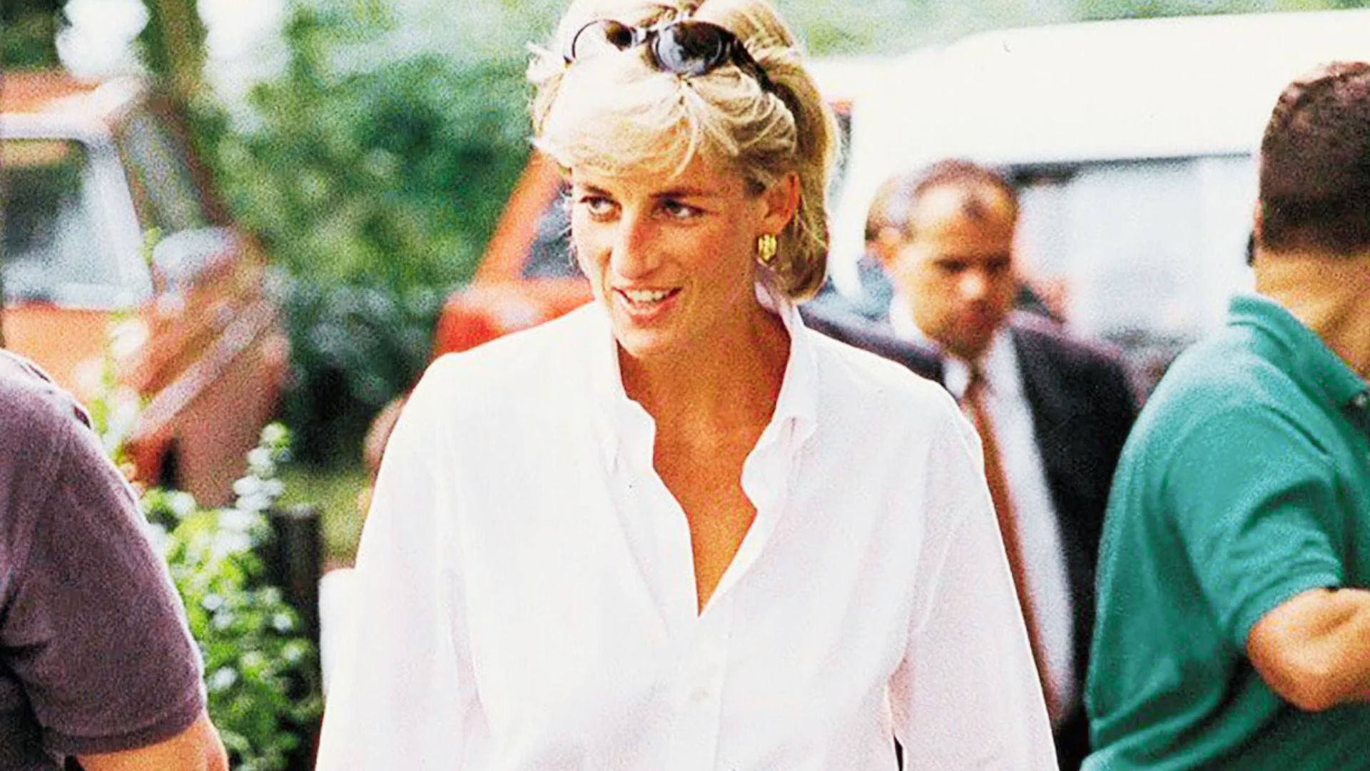 Princess Diana smiles while walking outdoors in a relaxed white button-down shirt with sunglasses pushed up on her head.