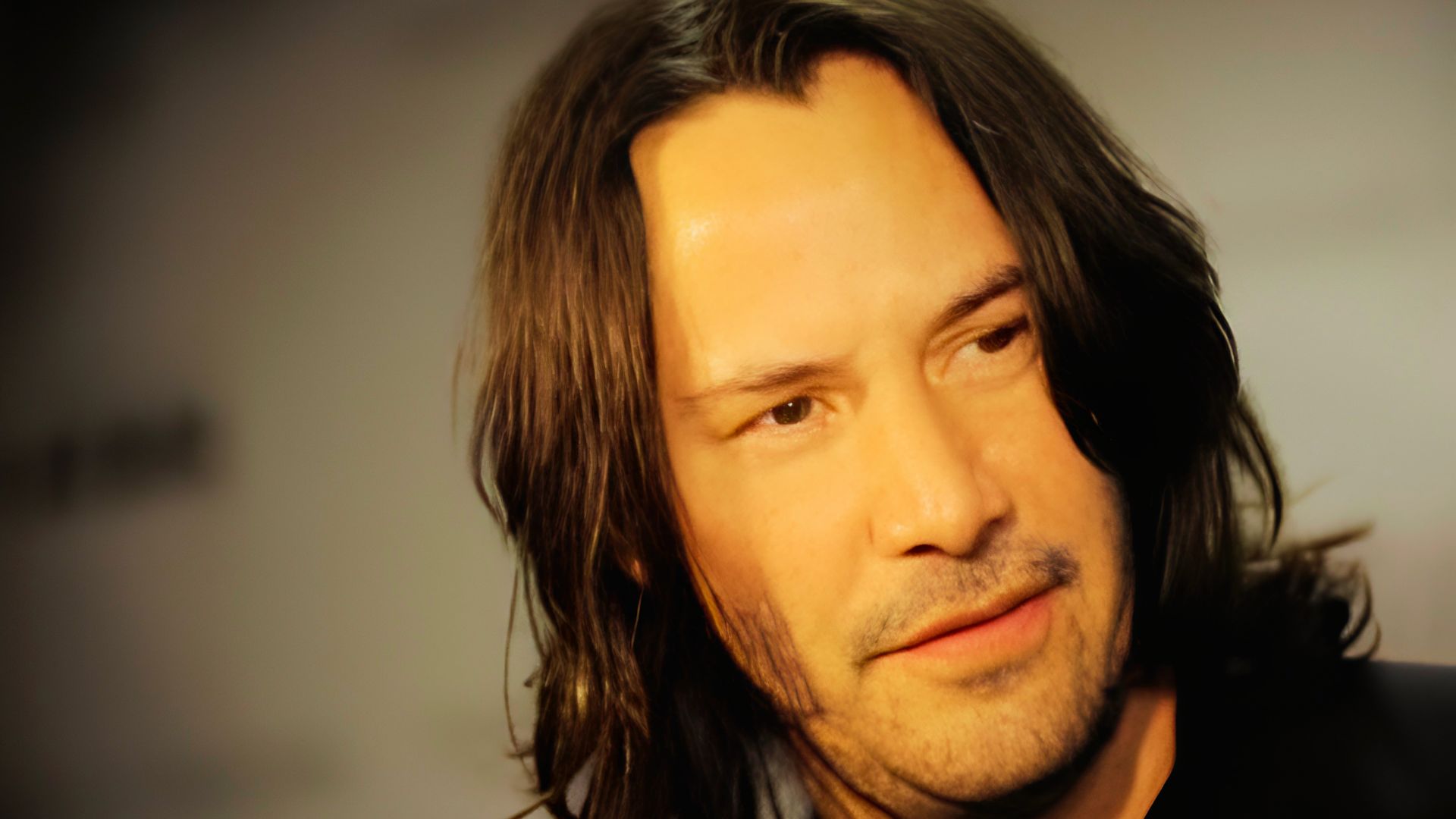 Young Keanu Reeves in a white t-shirt with short, dark, tousled hair, standing next to a surfboard.