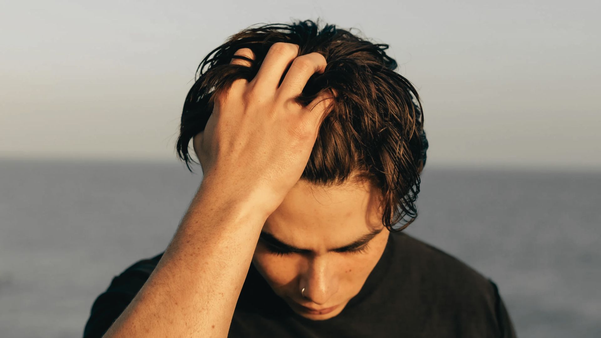 Young man running his hand through dark medium length wavy hair outdoors near the ocean in natural light.