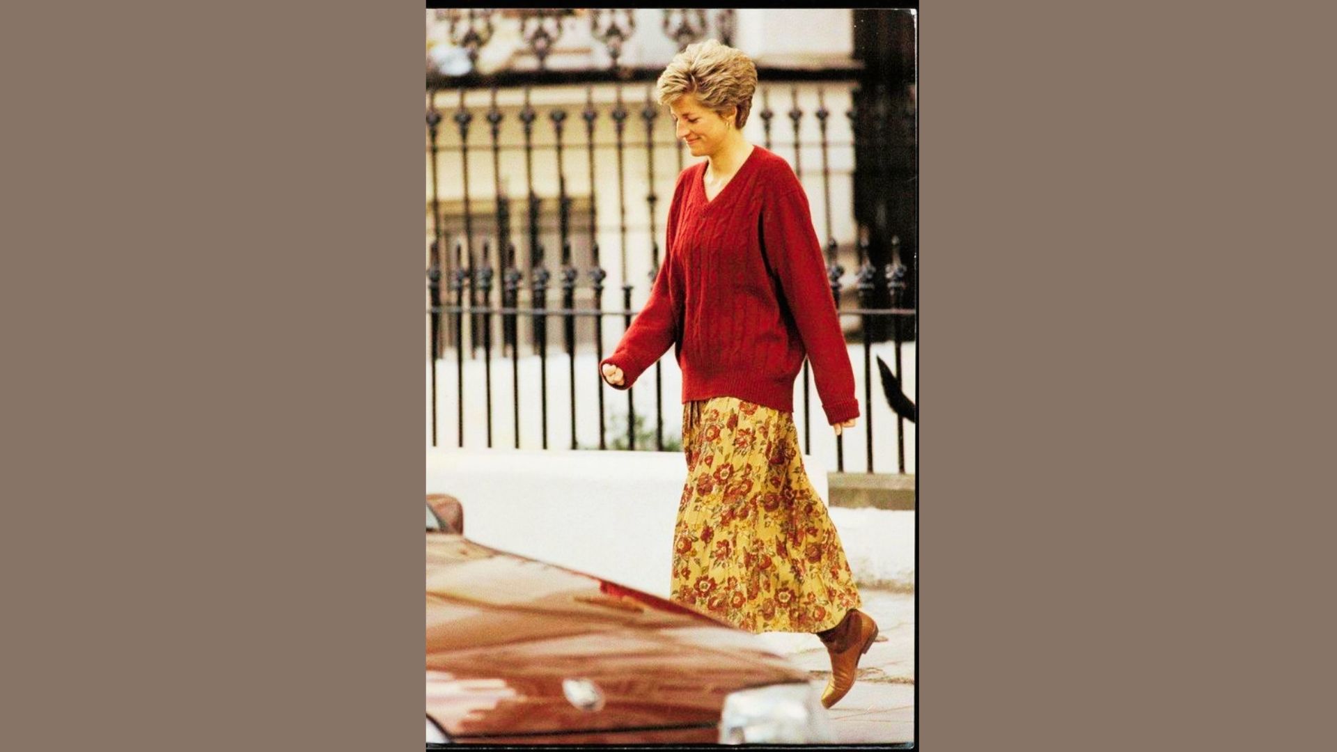 Princess Diana walks outdoors in a relaxed red cable-knit V-neck sweater paired with a long floral midi skirt.