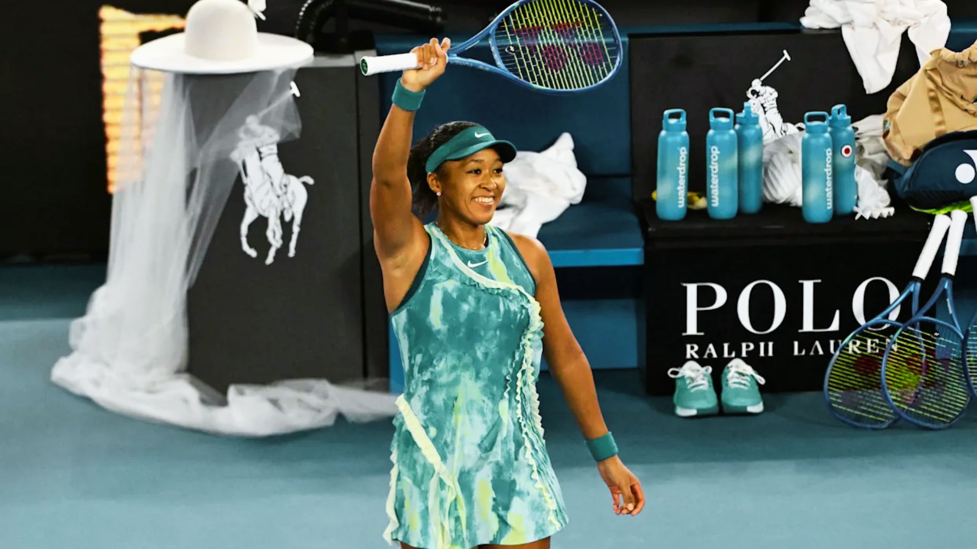 Naomi Osaka celebrates a win on court, raising her racket while wearing a teal tie-dye Nike dress with ruffle trim.
