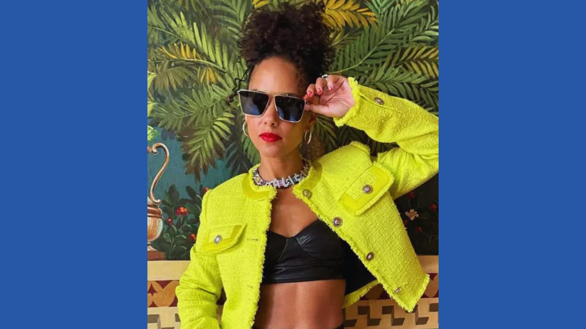 Alicia Keys in neon yellow tweed jacket wears curly high puff updo, oversized sunglasses, red lips, tropical mural background.