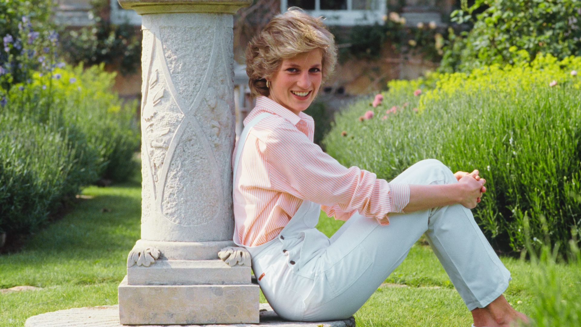 Princess Diana sits barefoot in a garden wearing white overalls over a pink striped button-down shirt.