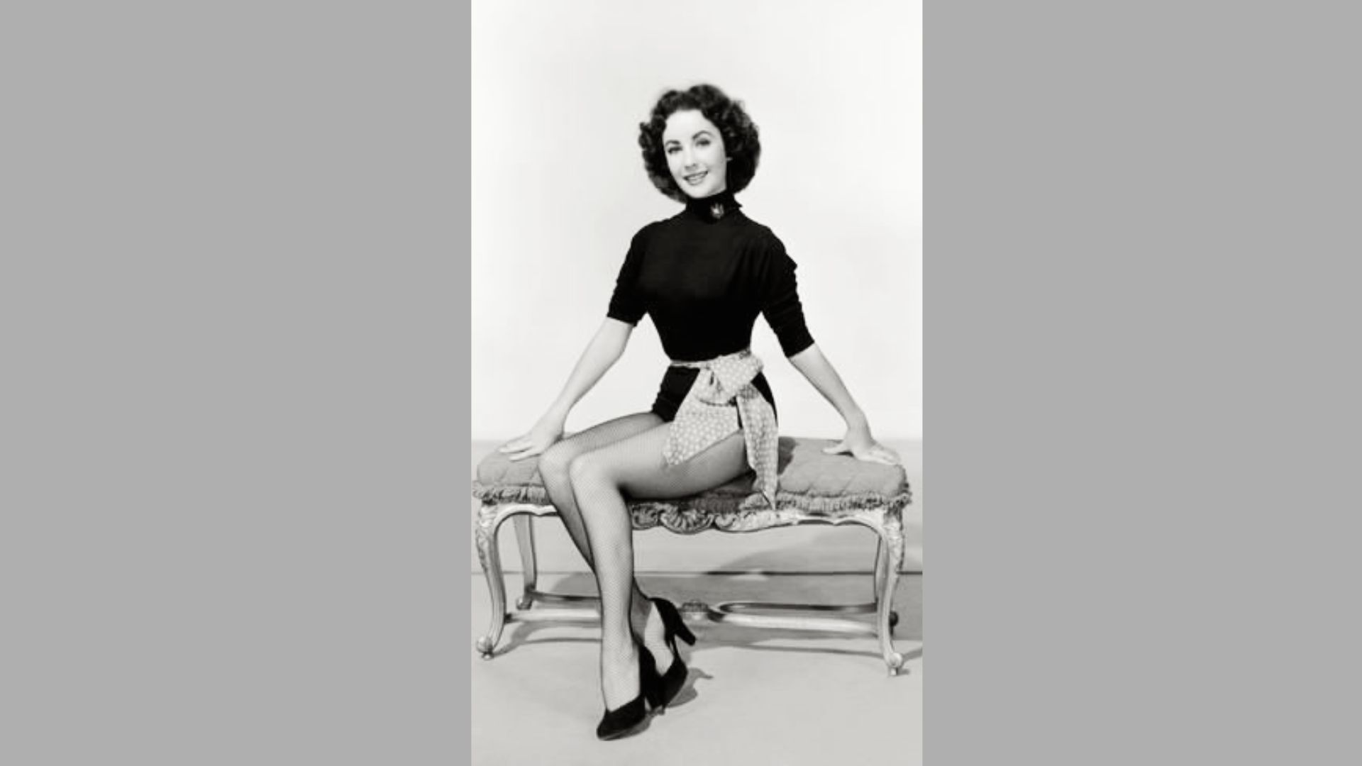 Elizabeth Taylor sits on an ornate bench in a black turtleneck leotard, fishnet stockings, and heels.