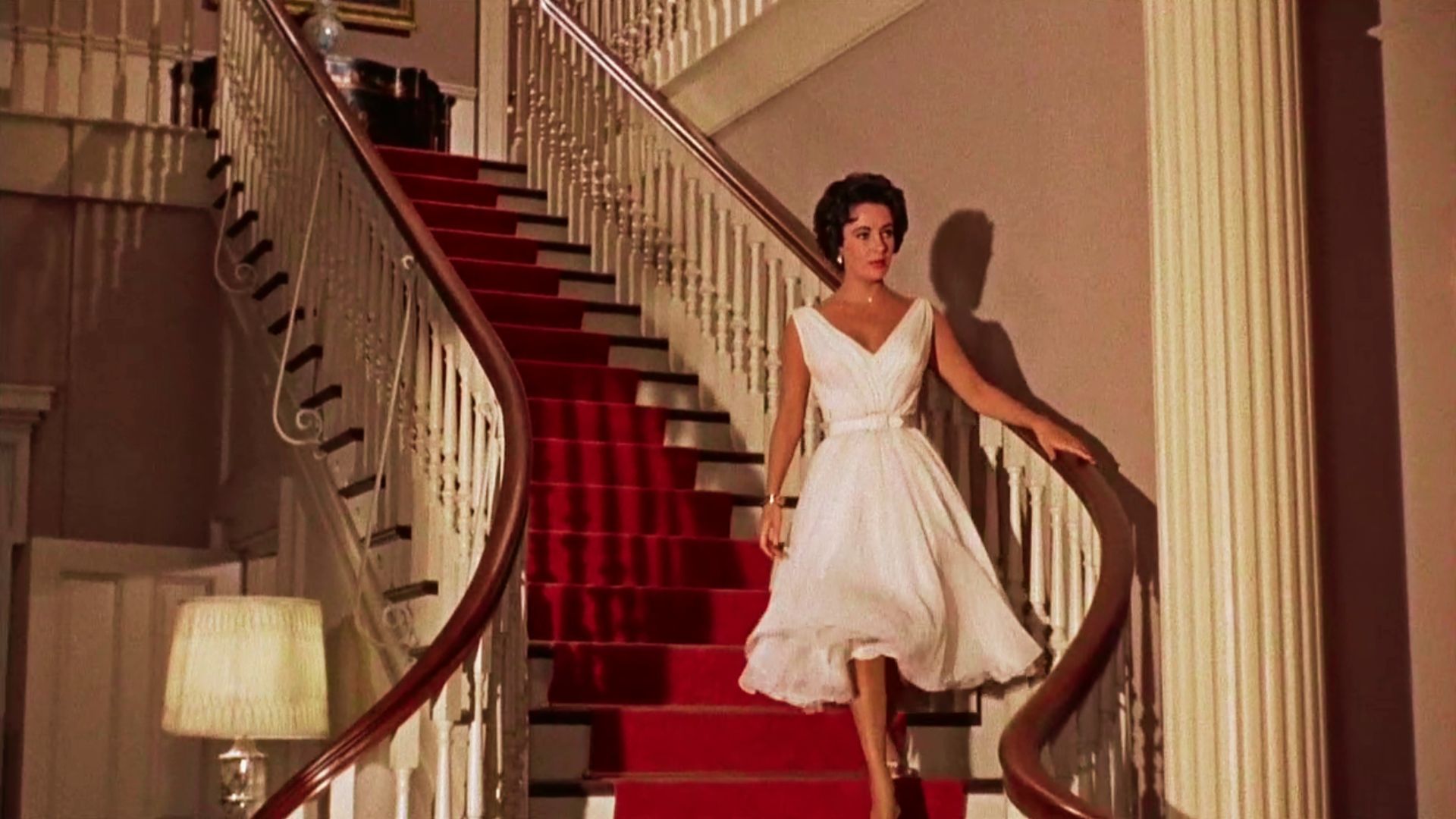 Elizabeth Taylor descends a red-carpeted staircase in a white V-neck midi dress with a full skirt.