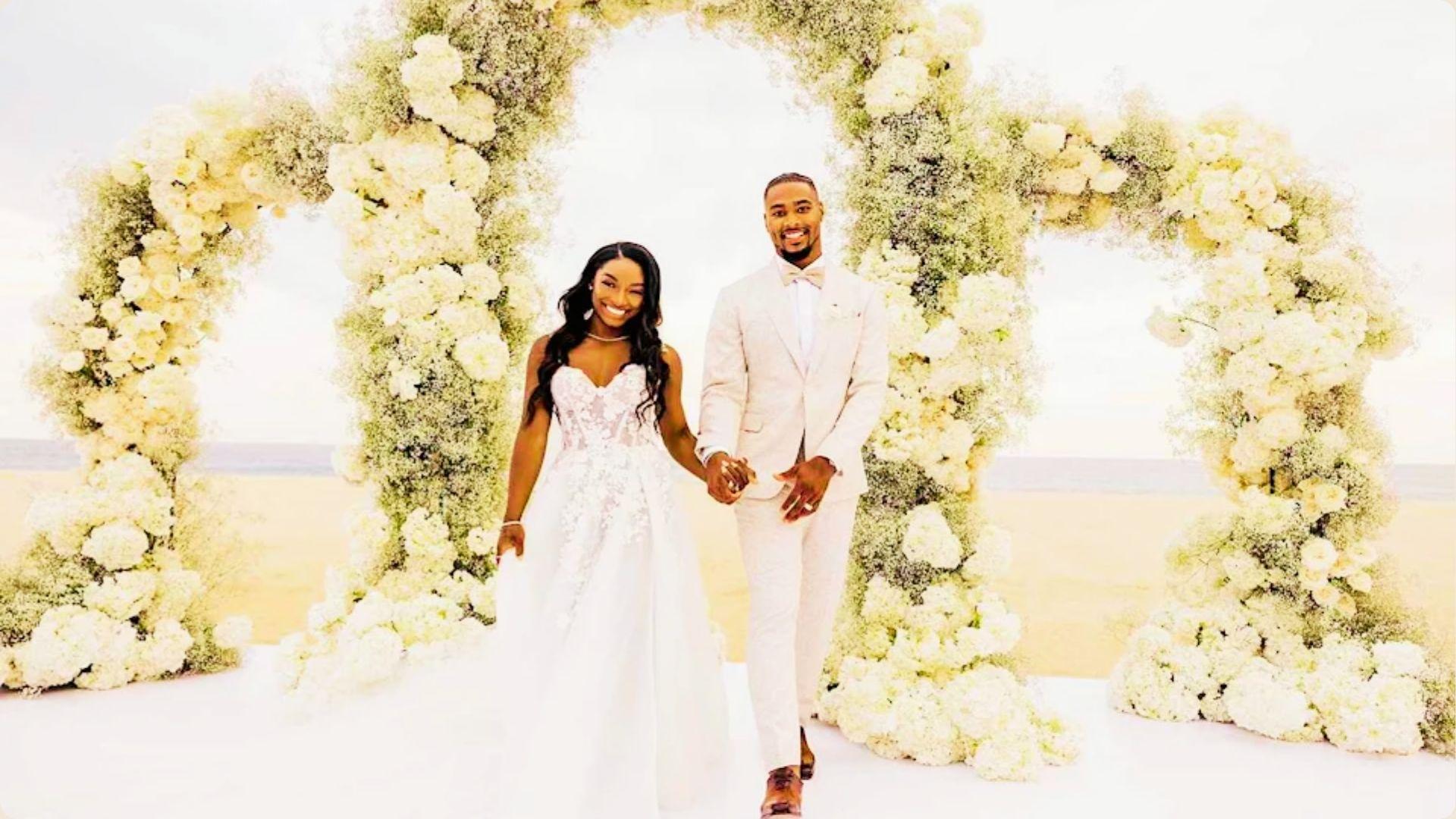 Simone Biles and Jonathan Owens hold hands under a large floral arch on the beach.