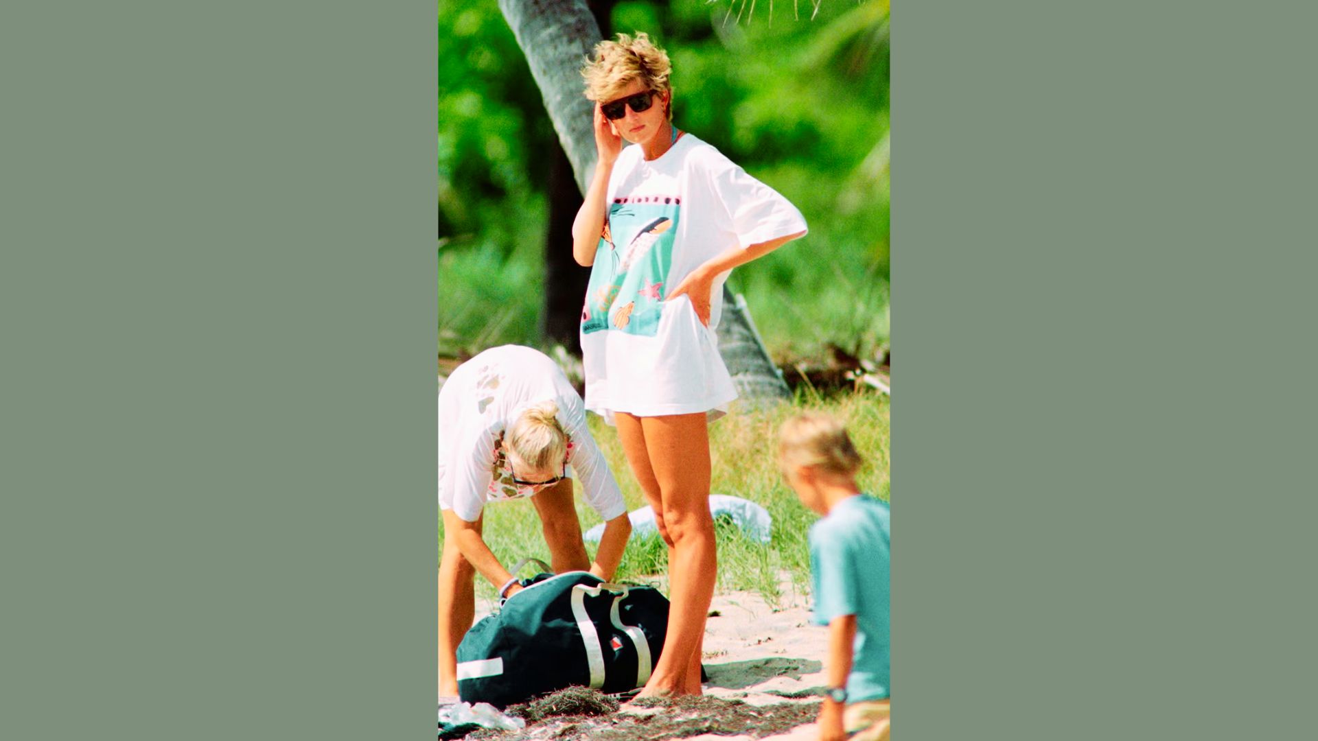 Princess Diana stands barefoot on a beach in an oversized white graphic T-shirt worn as a dress.