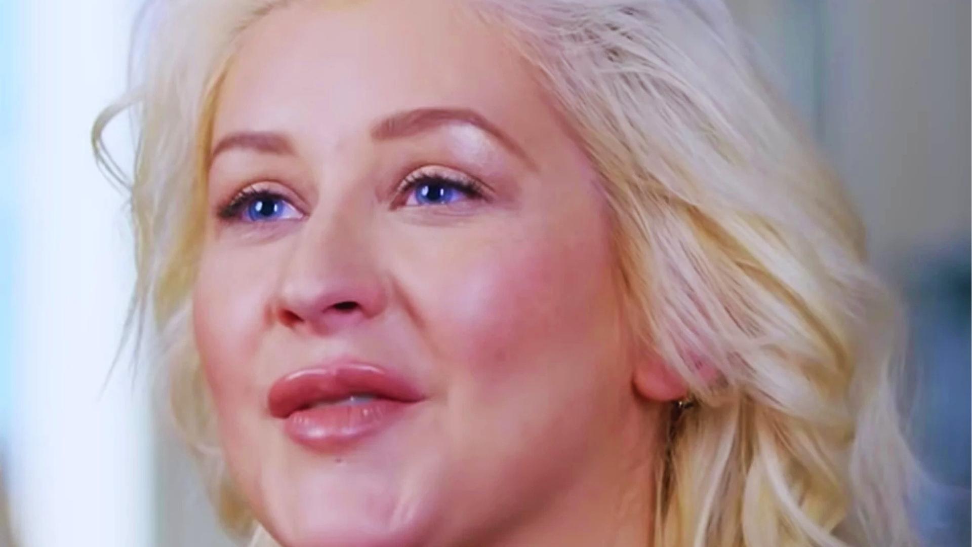 Christina Aguilera with soft blonde waves and minimal makeup looks upward, glossy lips and subtle blush visible in close-up.
