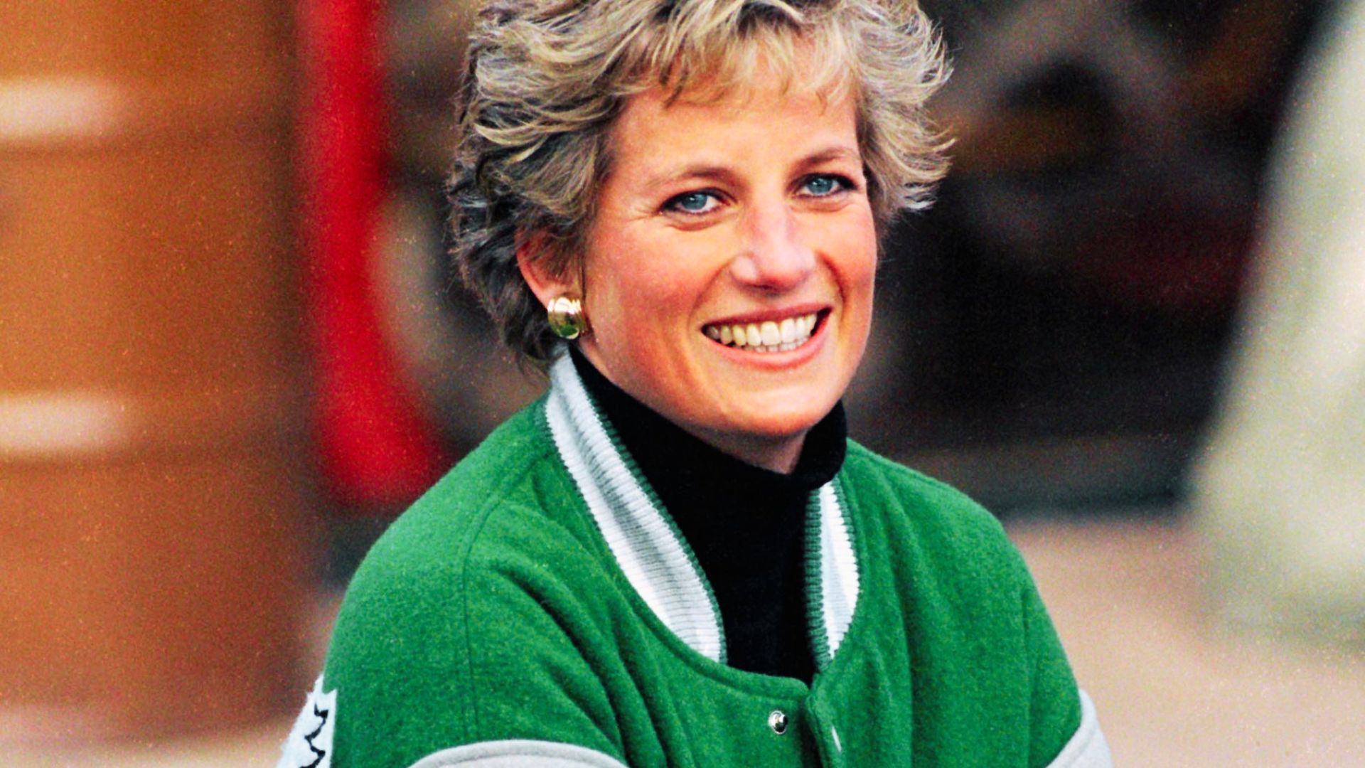 Princess Diana laughs outdoors in a green varsity jacket layered over a black turtleneck.
