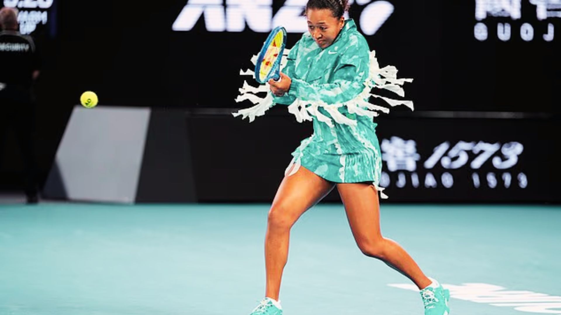 Naomi Osaka hits a backhand in a teal tie-dye Nike jacket with dramatic white fringe detailing at the Australian Open.