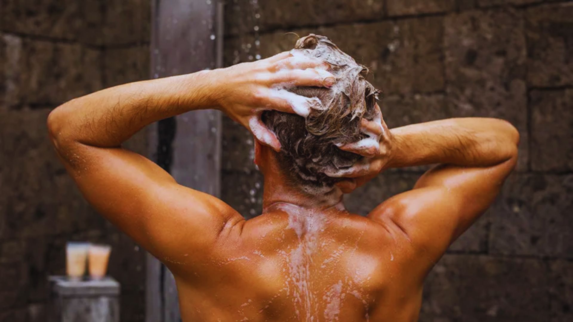 Man washing his hair with shampoo in an outdoor shower with stone walls in the background.