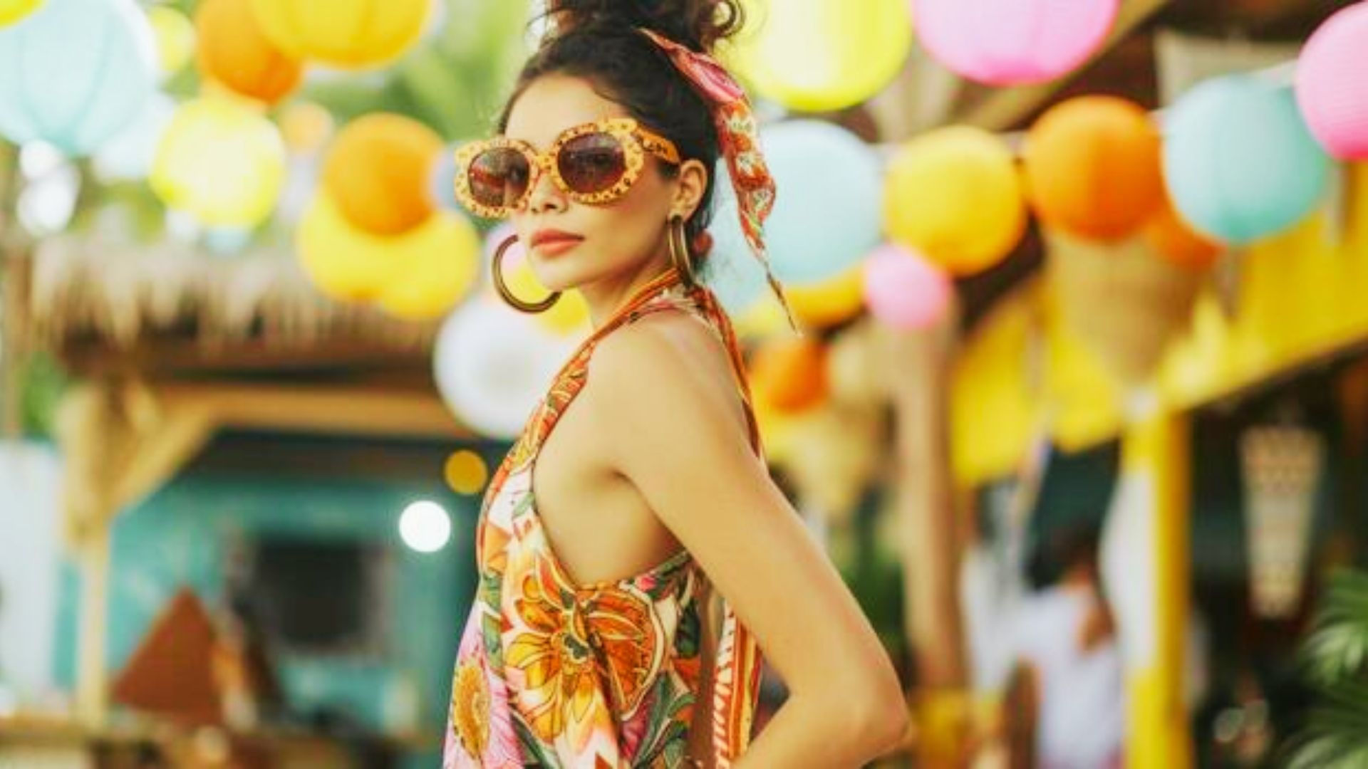 Woman in a vibrant floral halter dress with oversized sunglasses, hoop earrings, and a silk headscarf.