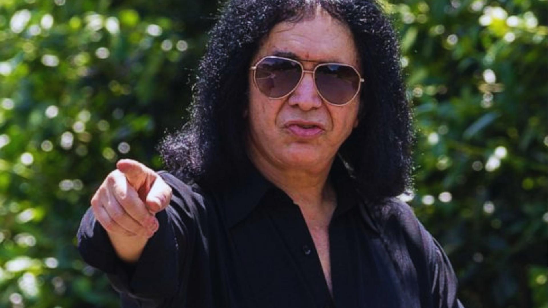 Gene Simmons points at the camera outdoors with no KISS makeup, aviator sunglasses, black shirt, and natural curly hair.