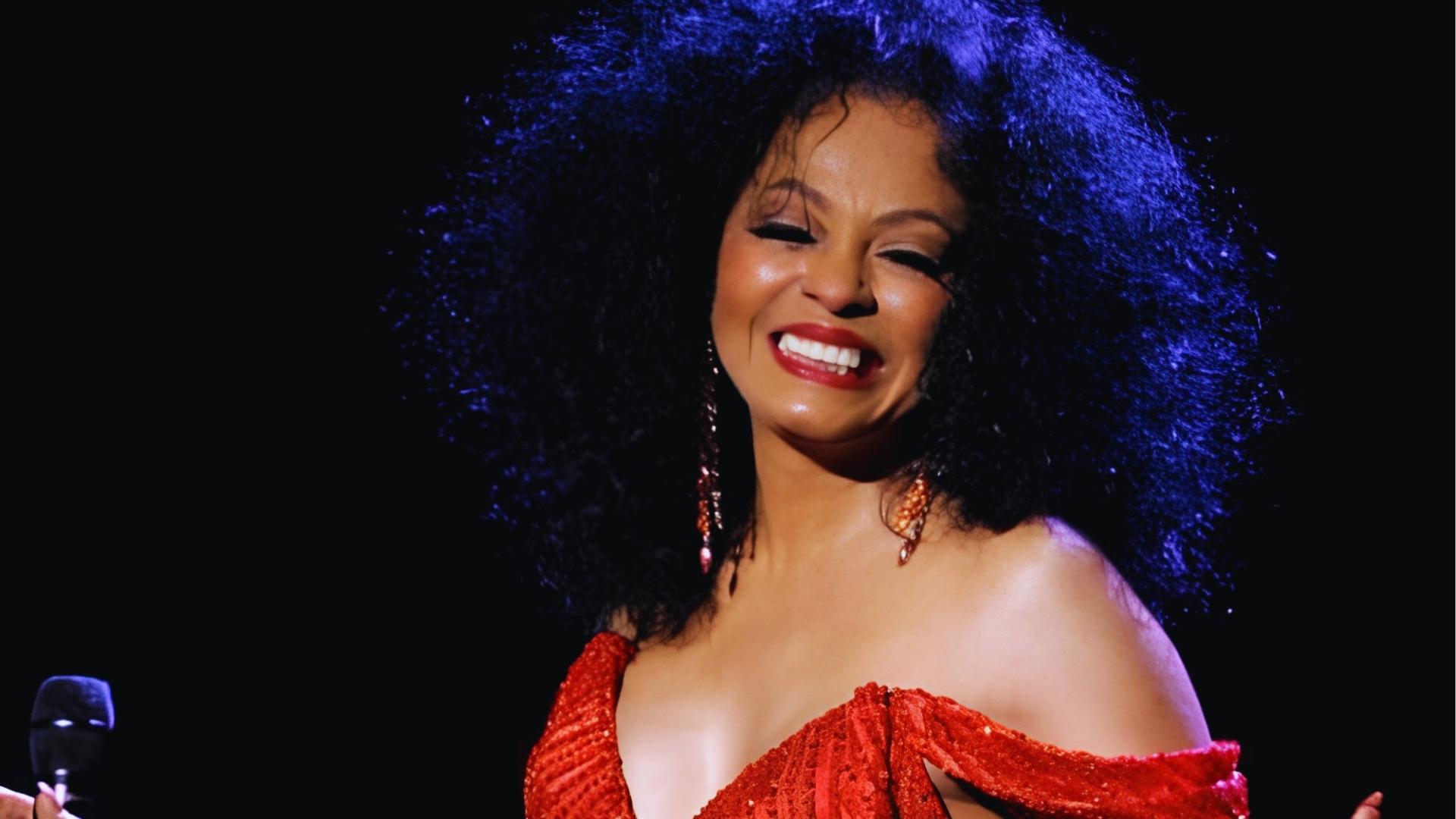 Diana Ross sings onstage in red off-shoulder gown, showcasing her iconic voluminous natural afro under dramatic blue stage lighting.