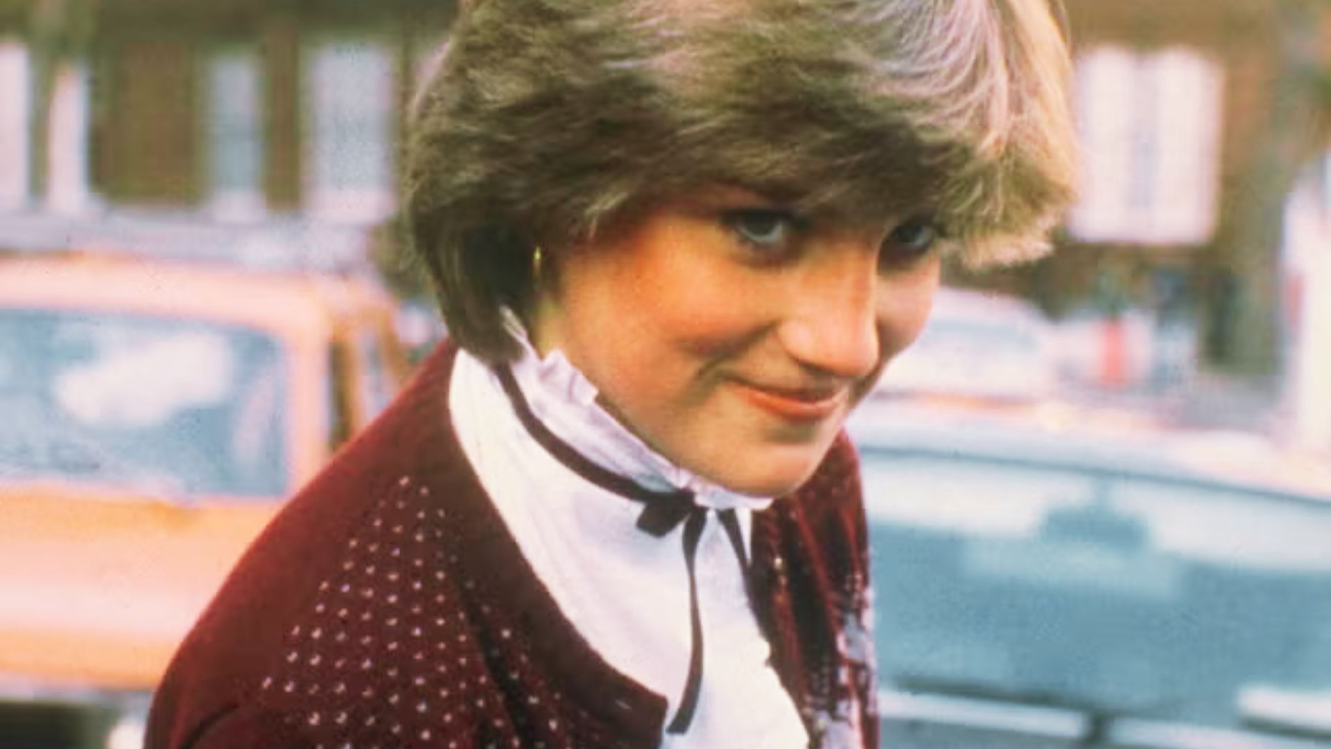 Princess Diana smiles shyly wearing a burgundy cardigan with dotted details over a white ruffled blouse with black ribbon tie.
