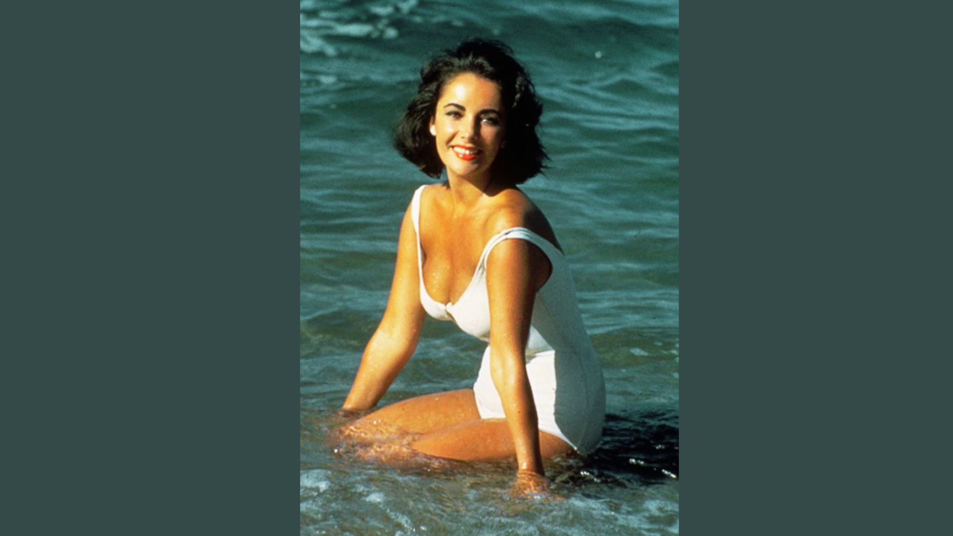 Elizabeth Taylor kneels in the surf in a white scoop-neck swimsuit, smiling at the camera.