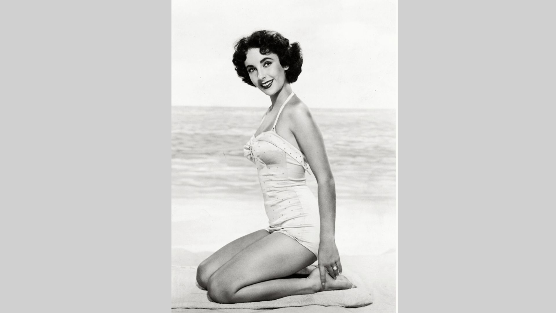 Young Elizabeth Taylor kneels on a beach in a white halter swimsuit with polka-dot detail.