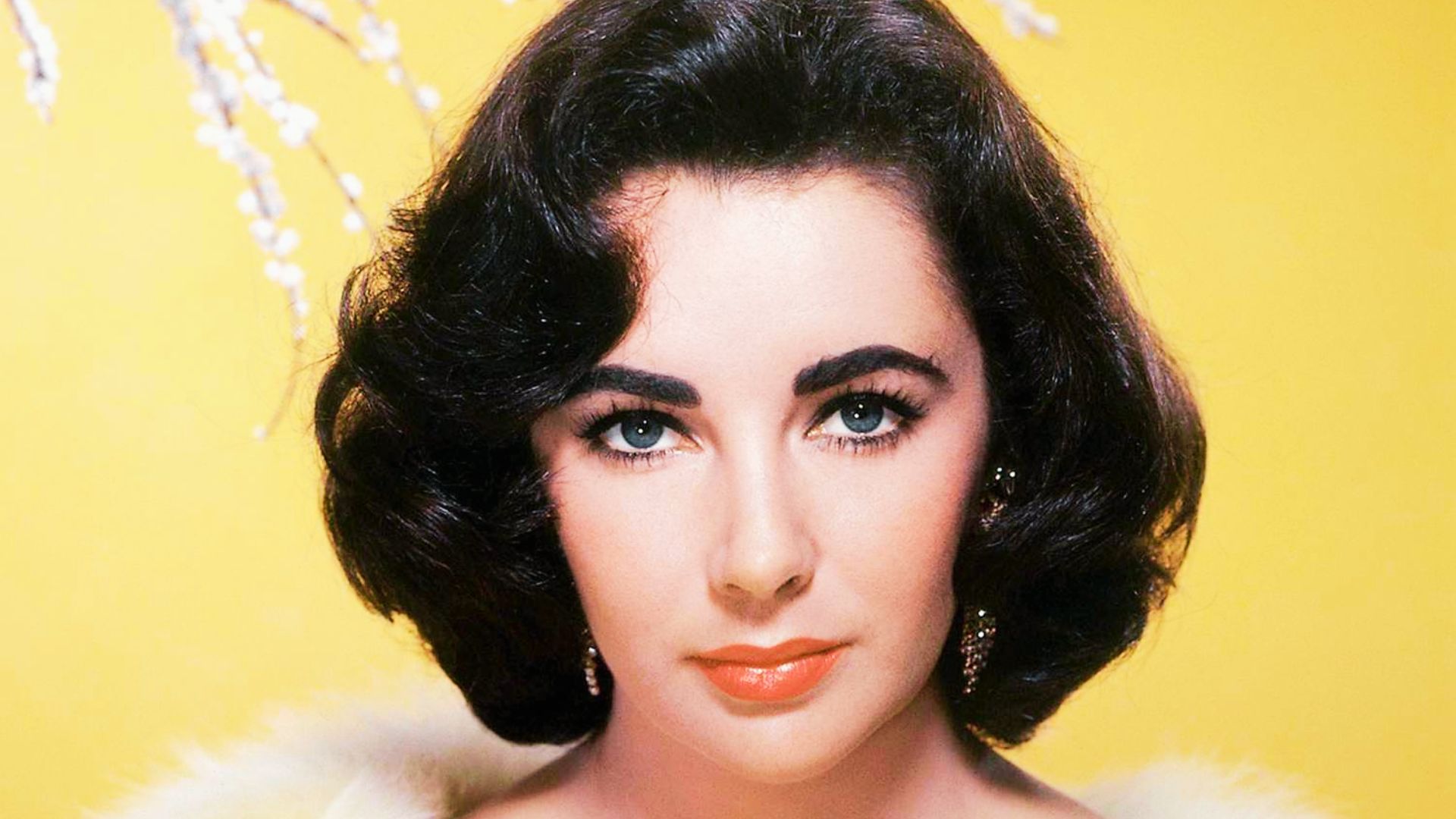 Elizabeth Taylor poses against a bright yellow backdrop with voluminous dark curls, coral lips, and gold drop earrings.