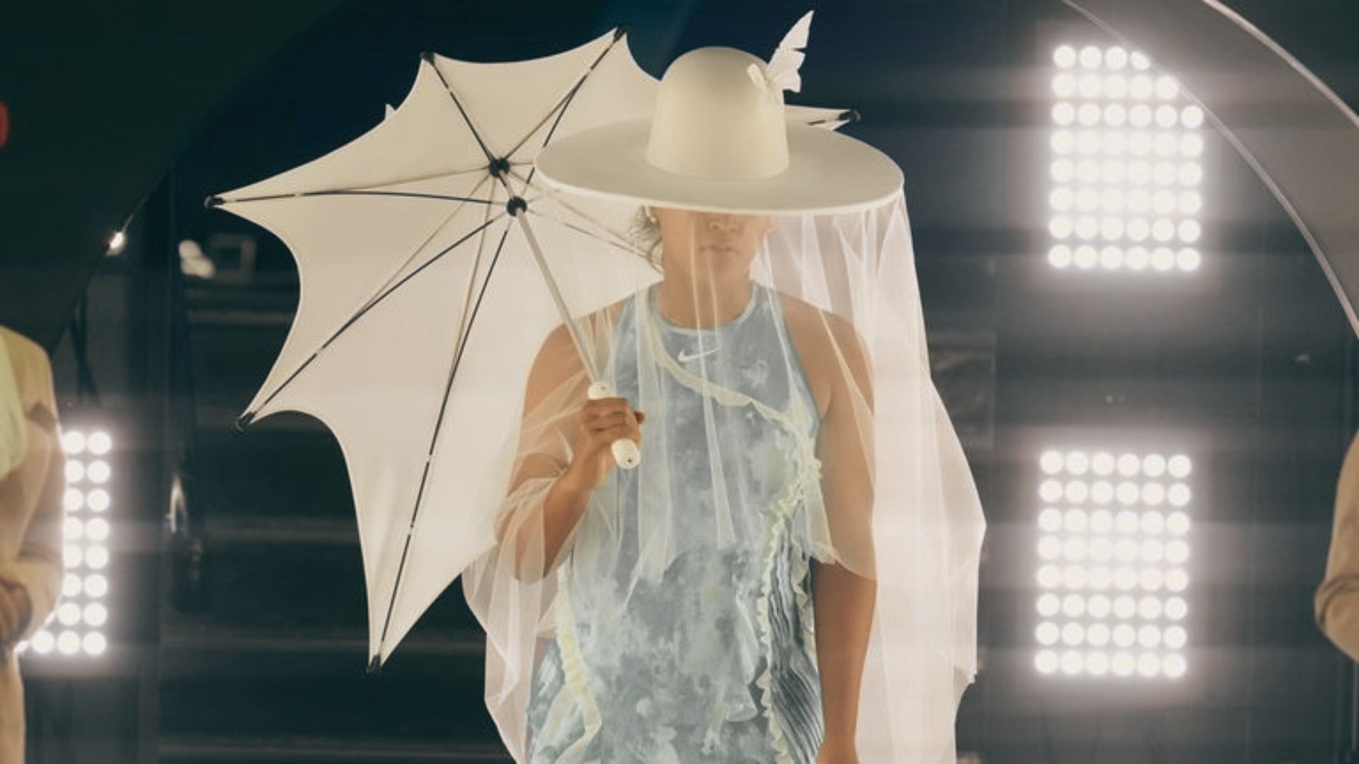 Naomi Osaka walks onto the court holding a white parasol, wearing a wide-brimmed white hat with a sheer veil and butterfly detail.
