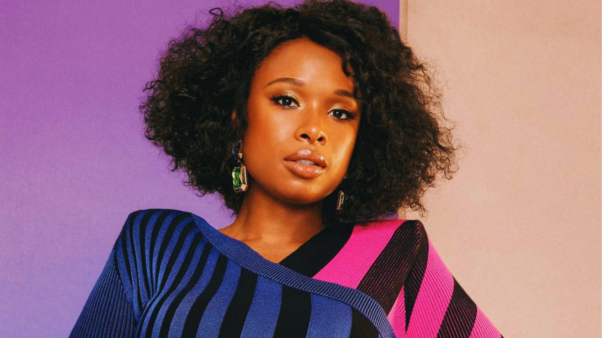 Jennifer Hudson wears blue and pink striped sweater with rounded natural afro, posing against a purple and beige split background.