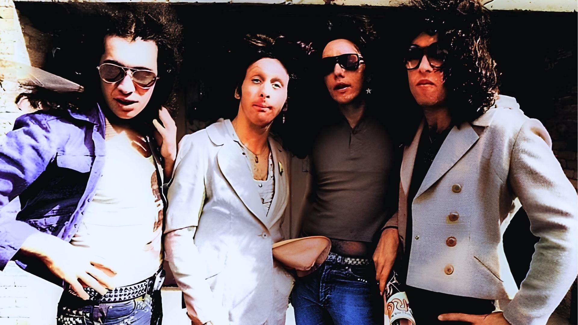 Colorized 1970s photo of KISS without makeup, Gene Simmons far right in white blazer, sunglasses, and voluminous curly hair.