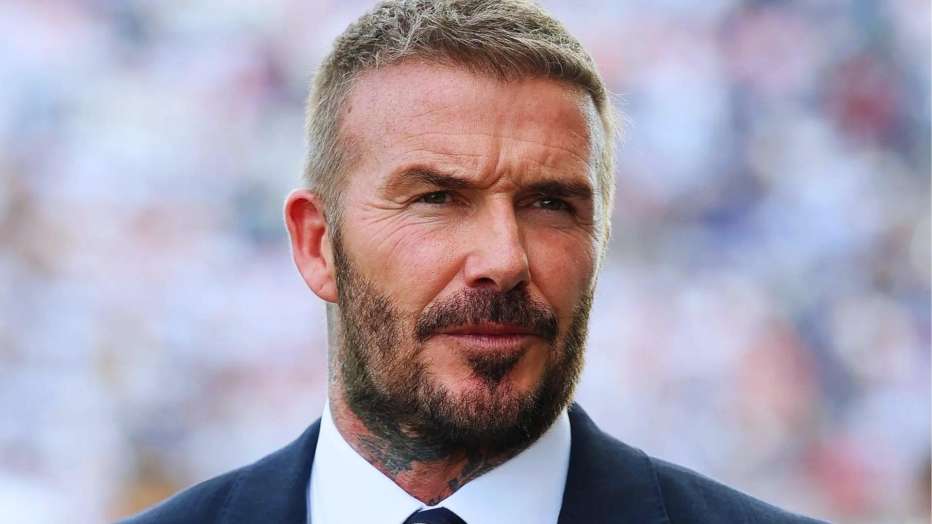 David Beckham at stadium with short salt-and-pepper hair, fuller appearance, trimmed beard, wearing dark suit.