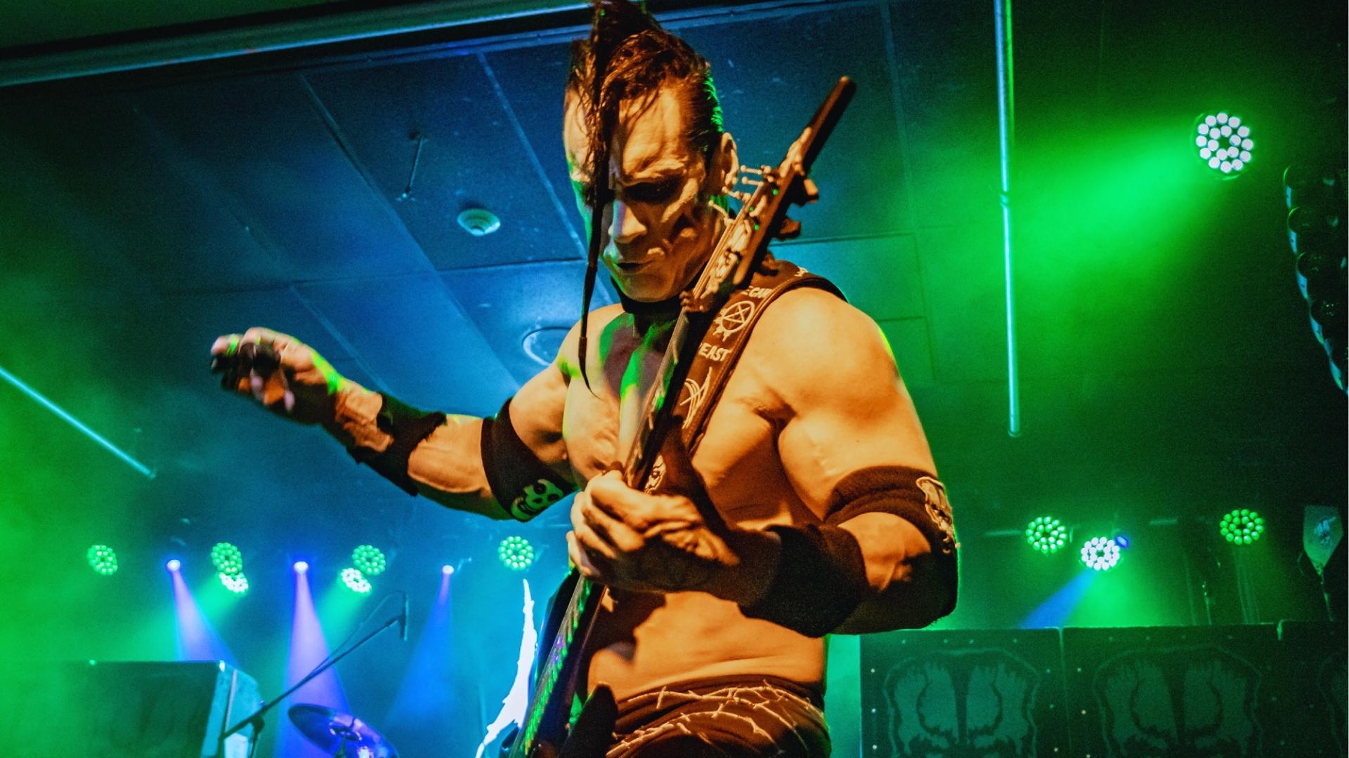 Doyle Wolfgang von Frankenstein shirtless onstage plays guitar with signature devilock, white face paint, black armbands under green lights.