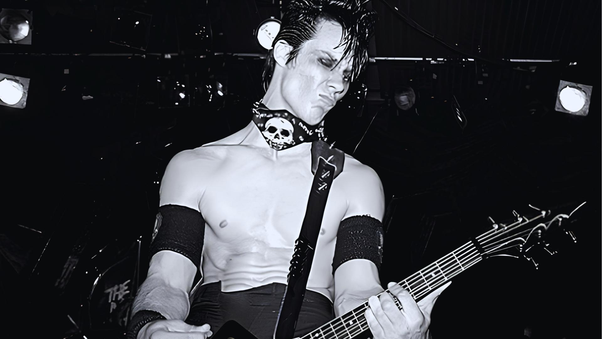 Black and white photo of young shirtless Doyle performing live with slicked devilock, skull bandana, and black armbands on guitar.