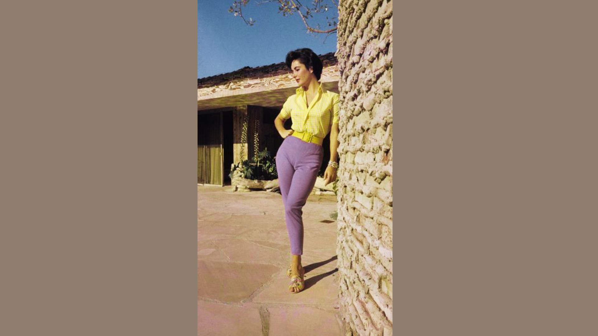 Elizabeth Taylor leans against a stone wall in a yellow gingham blouse and lavender capri pants.