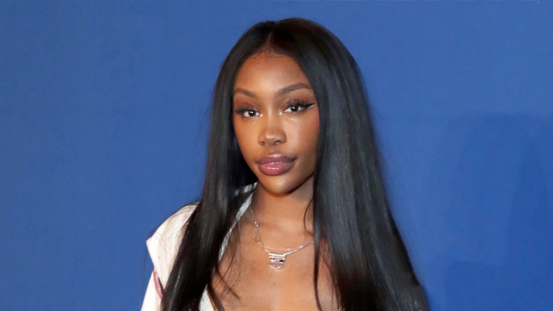 SZA with long, sleek straight black hair wearing a white top, posing confidently against a neutral background.