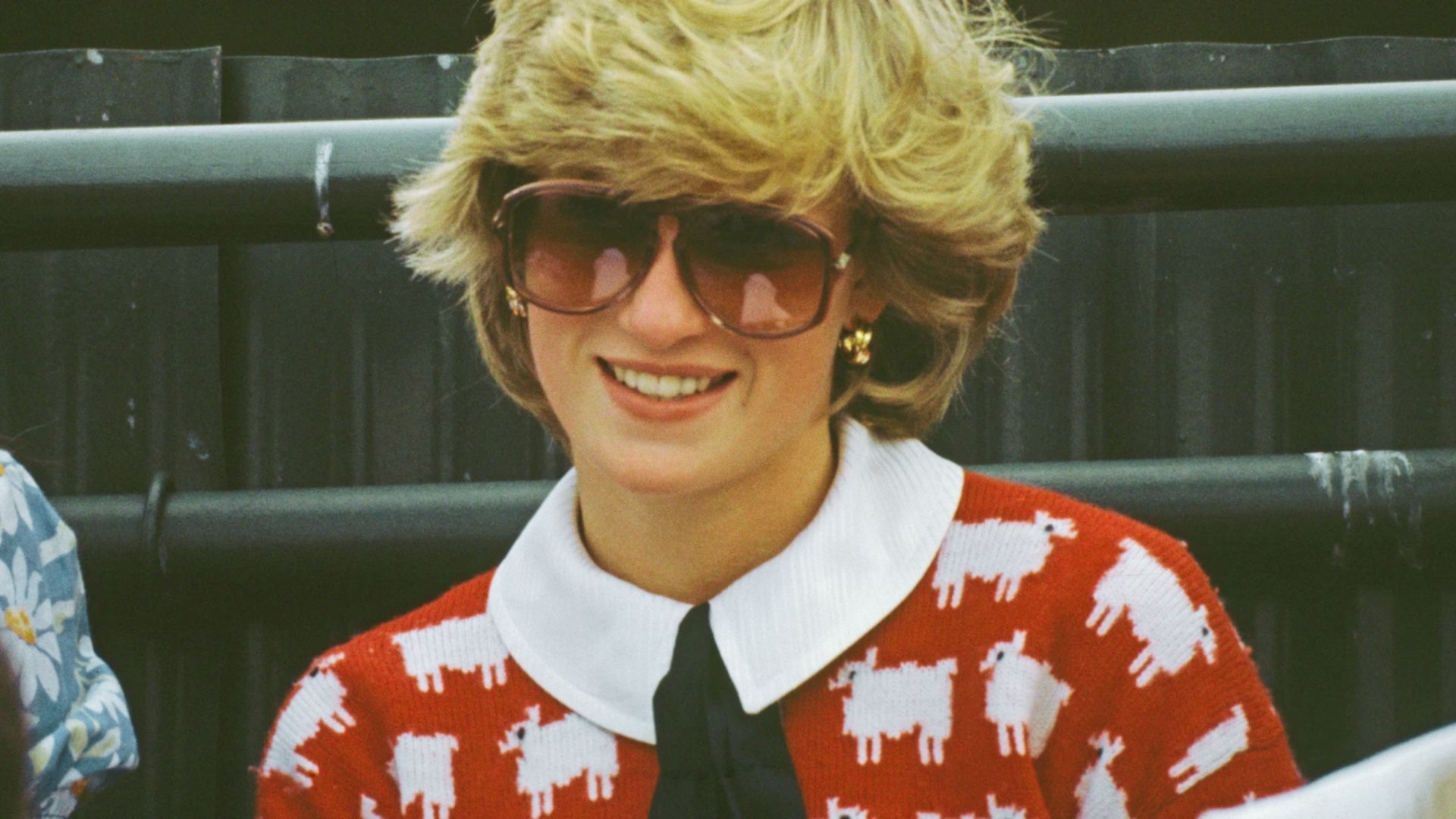 Princess Diana grins in her iconic red "black sheep" sweater layered over a white collared shirt with black tie.