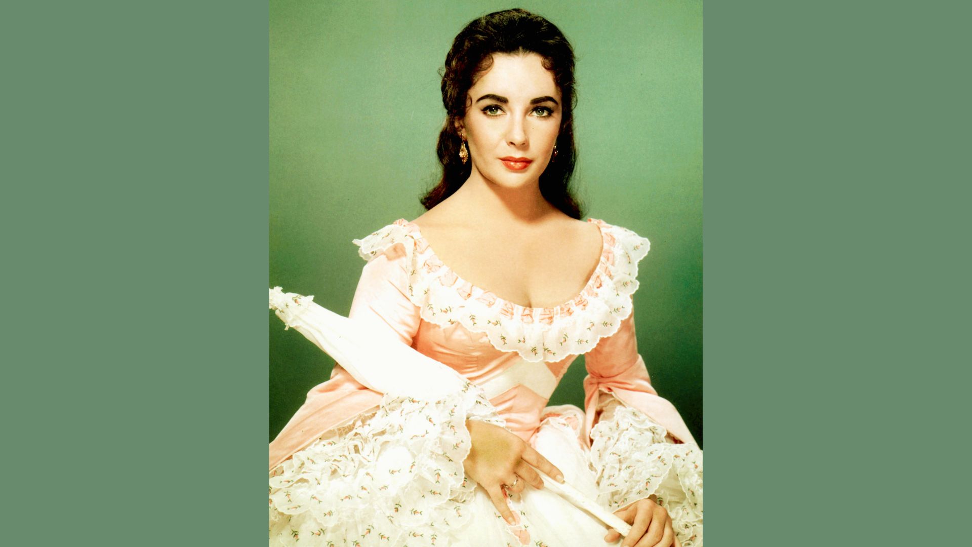 Elizabeth Taylor in a blush pink period costume with ruffled lace trim, holding a parasol.