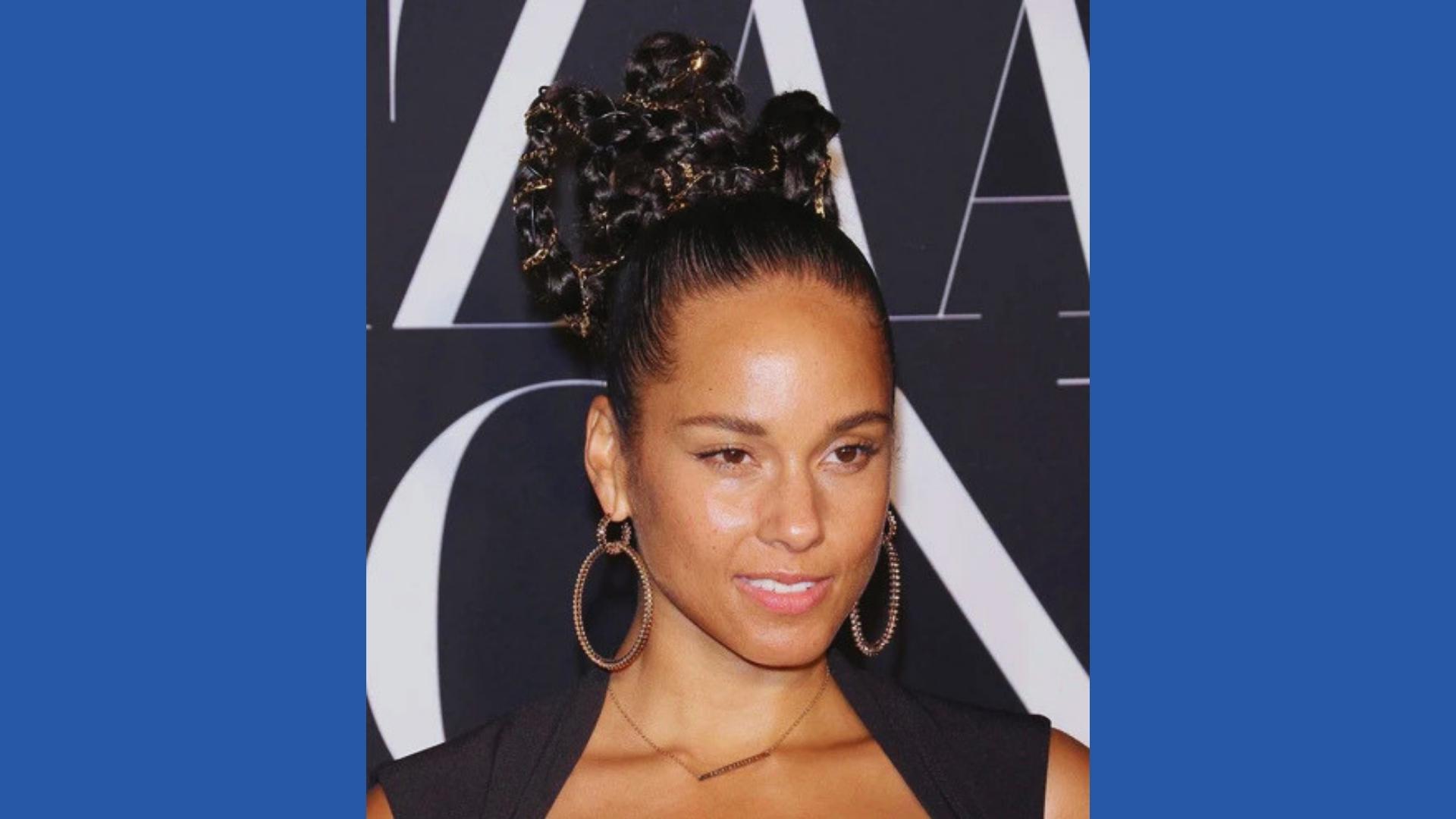 Alicia Keys at Harper’s Bazaar with sculptural braided updo woven with gold chains, sleek edges, black dress, large hoops.