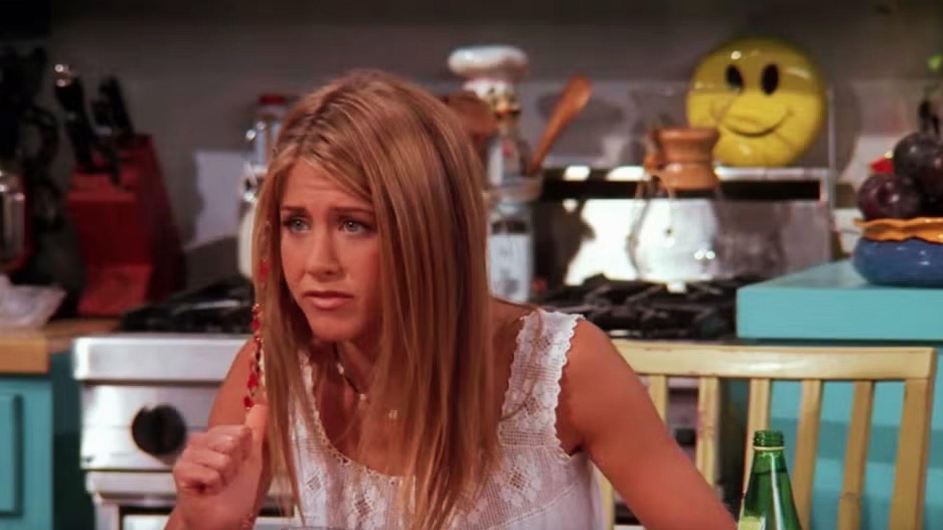 Rachel Green with long straight highlighted hair and beaded braid detail, wearing white lace top, sits in Monica’s kitchen.