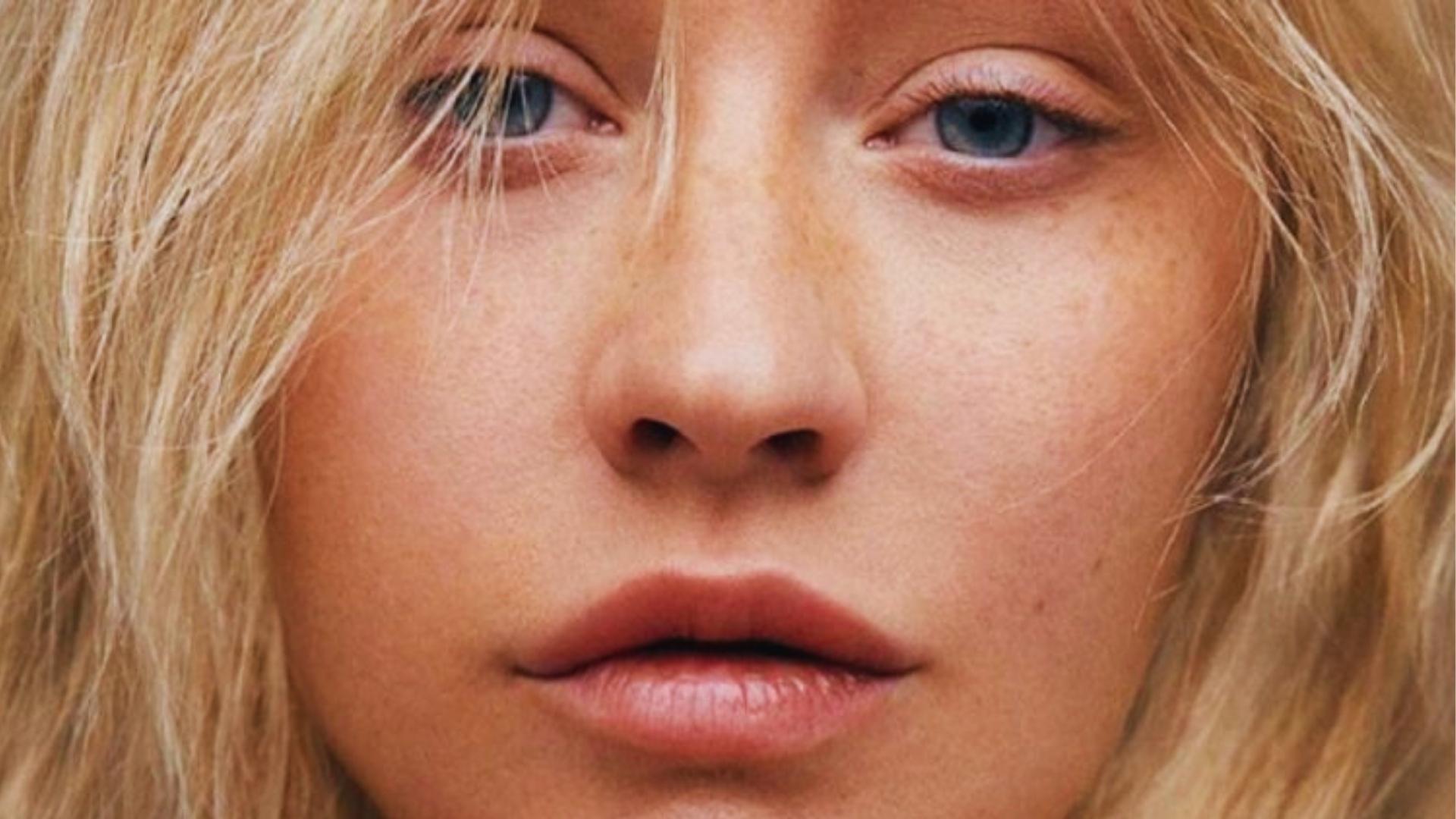 Extreme close-up of Christina Aguilera showing freckled bare skin, blue eyes, tousled blonde hair, and nude glossy lips.