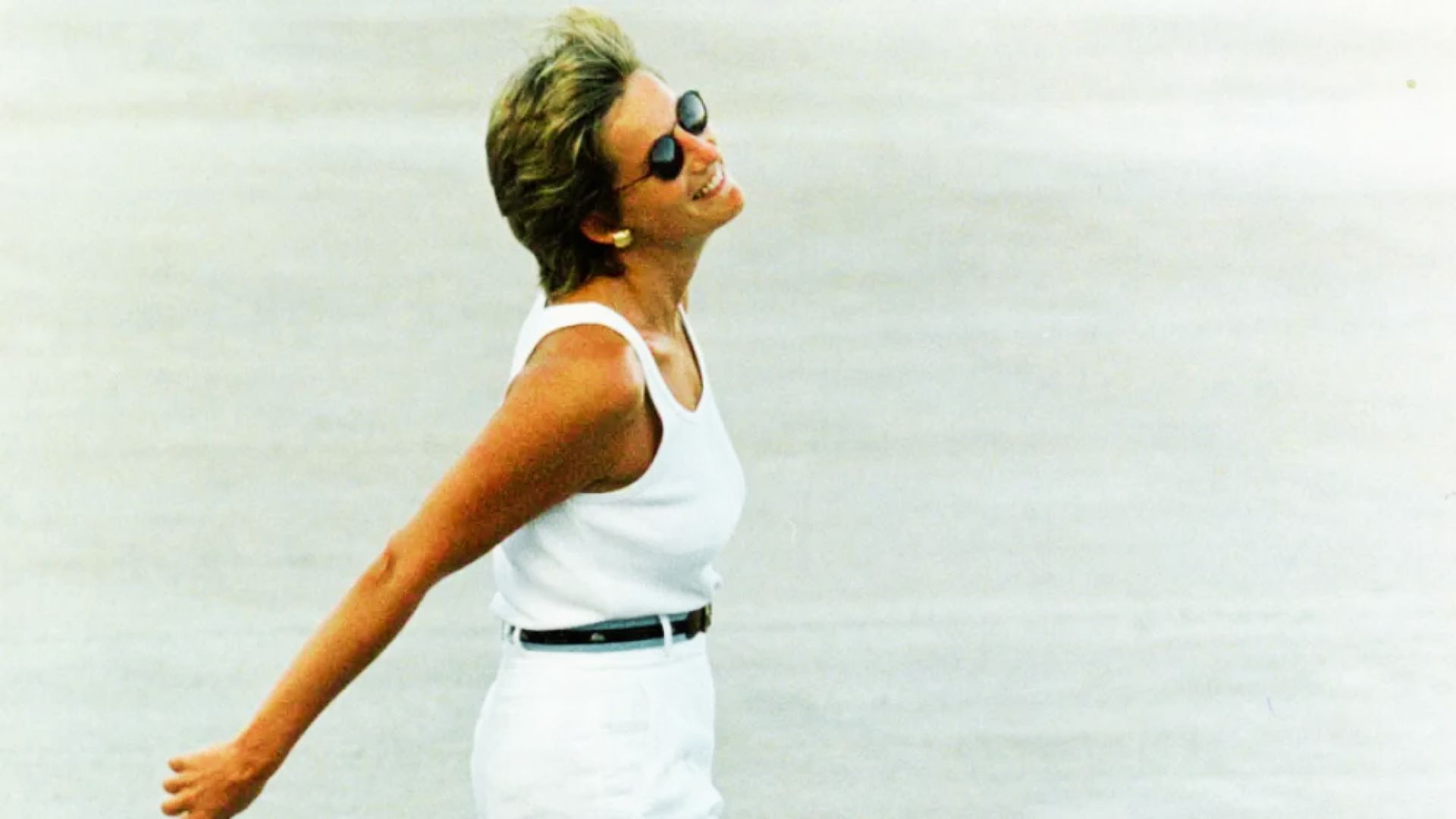 Princess Diana smiles in profile wearing an all-white outfit with a sleeveless top, high-waisted pants, and a thin black belt.