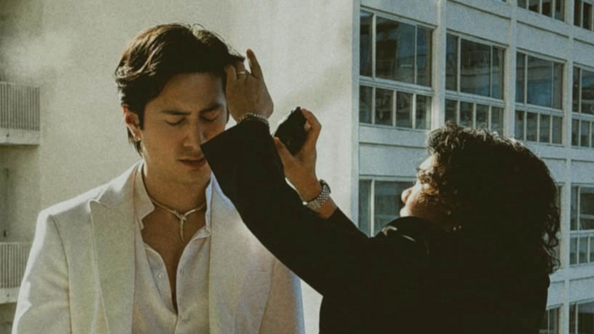 Hudson Williams getting his dark wavy hair styled by a hairstylist outdoors while wearing a white blazer.