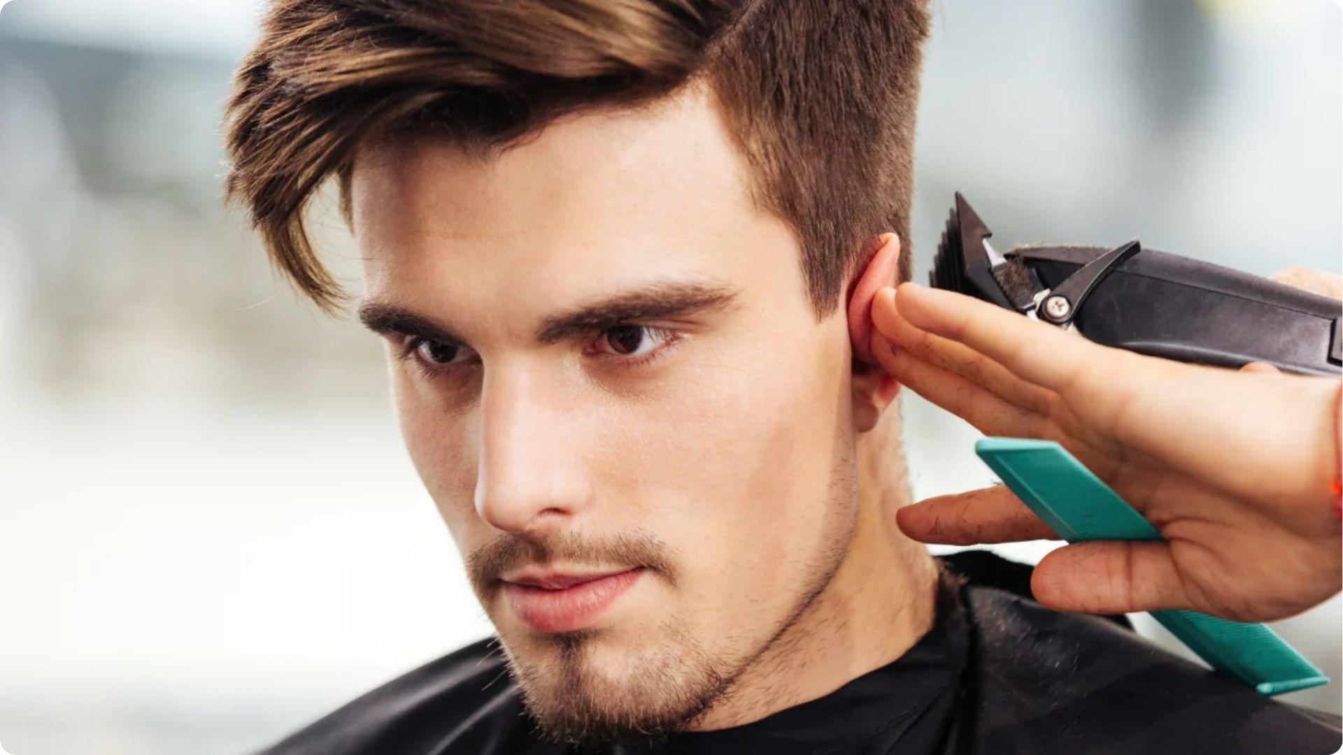 Barber doing clipper fade around ear on young man with side swept short hair, modern style