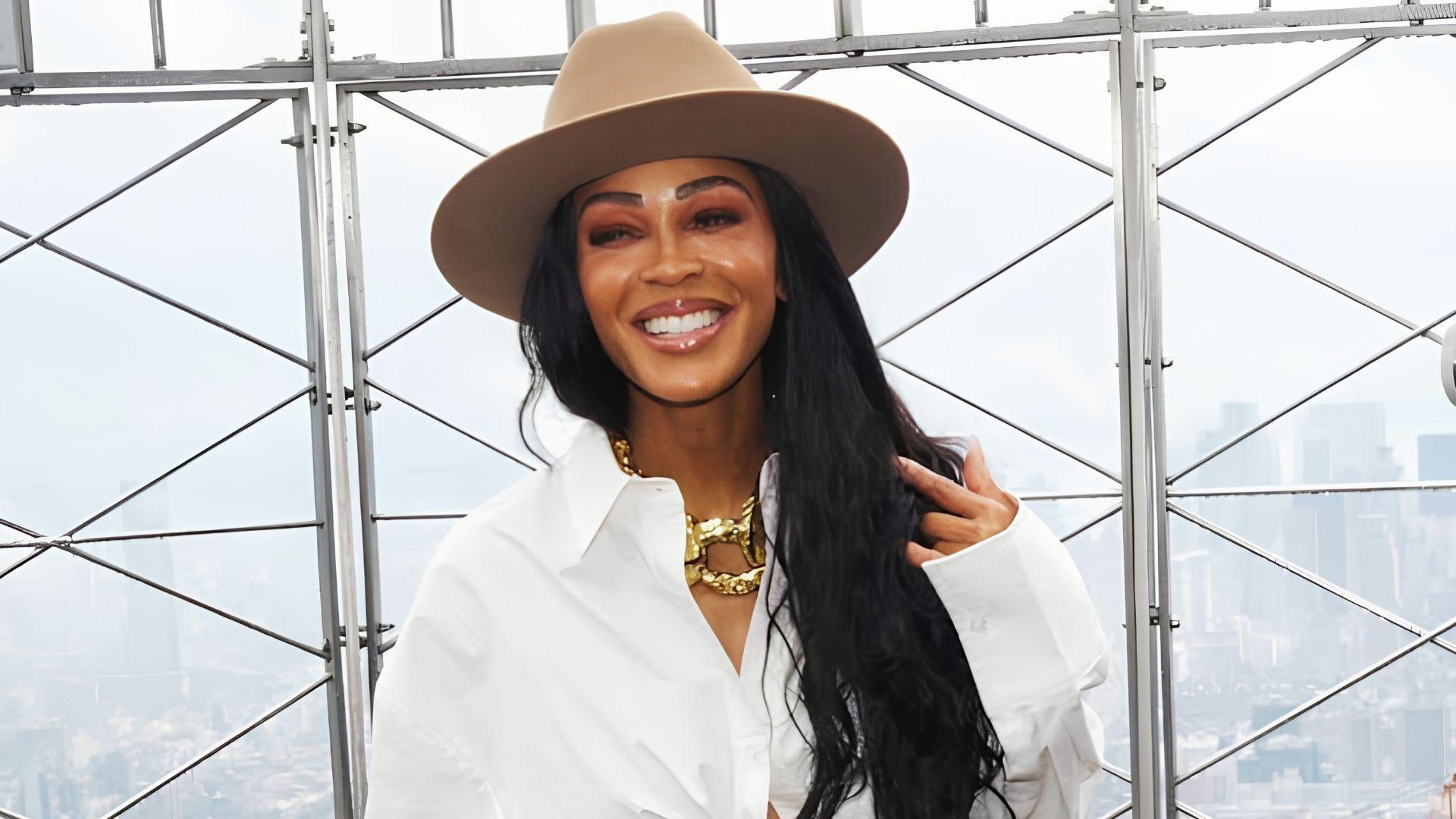 Meagan Good with long black wavy hair wearing a beige fedora hat and white shirt