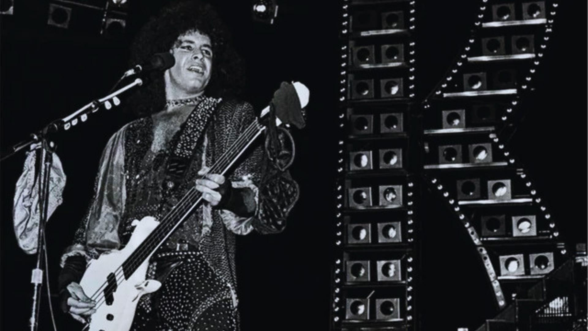Vintage black and white photo of young Gene Simmons without makeup playing white bass beside stacked KISS amplifiers onstage.